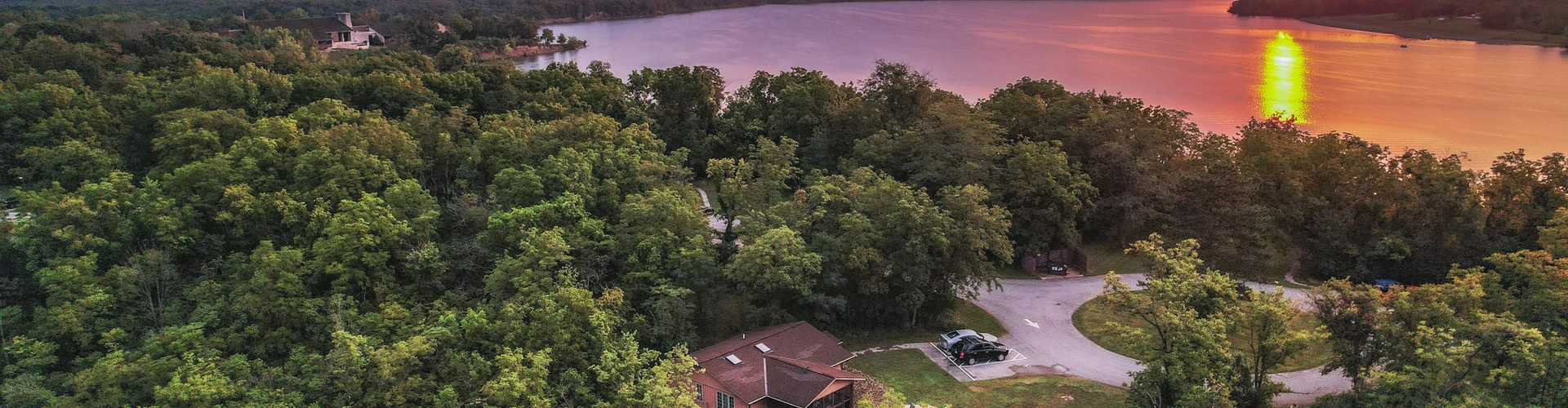 Cabin sits in a wooded area with a sunset over a lake.