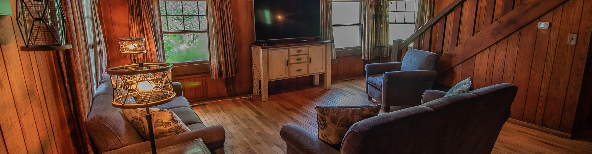 Cozy living room inside the Historic Harding Cabin featuring wood-paneled walls, vaulted ceilings, comfortable seating, warm lighting, rustic furnishings, and a staircase leading to the upper level.