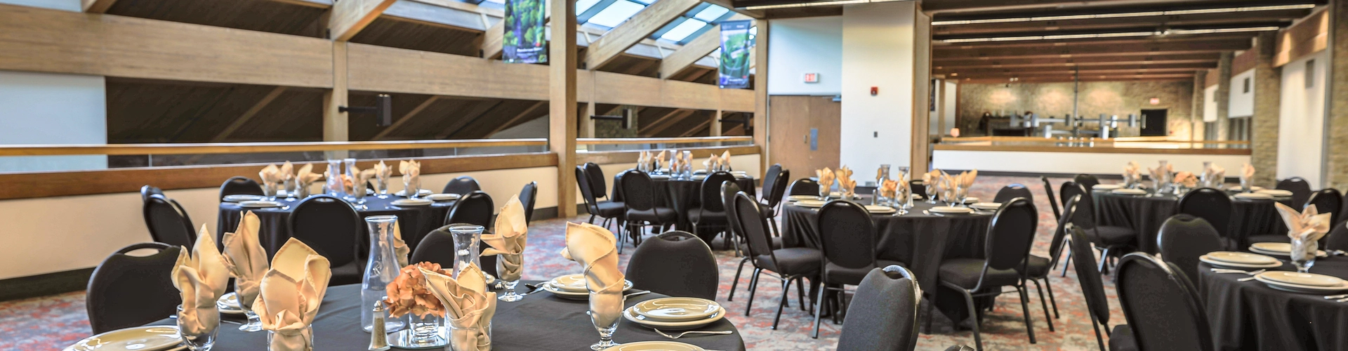 The deer creek mezzanine filled with large round tables covered by black table cloths surrounded by black cushioned chairs. Each seat is set with plates, utensils, drinking goblets, and cream-colored napkins