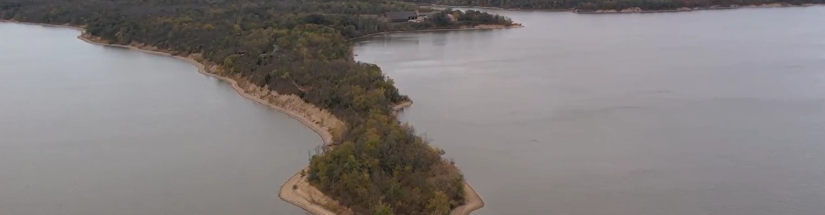 An aerial drone view of a narrow, densely forested peninsula extending out into the calm waters of the Deer Creek reservoir.